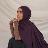 Woman wearing a dark purple hijab and brown long-sleeve top against a beige background