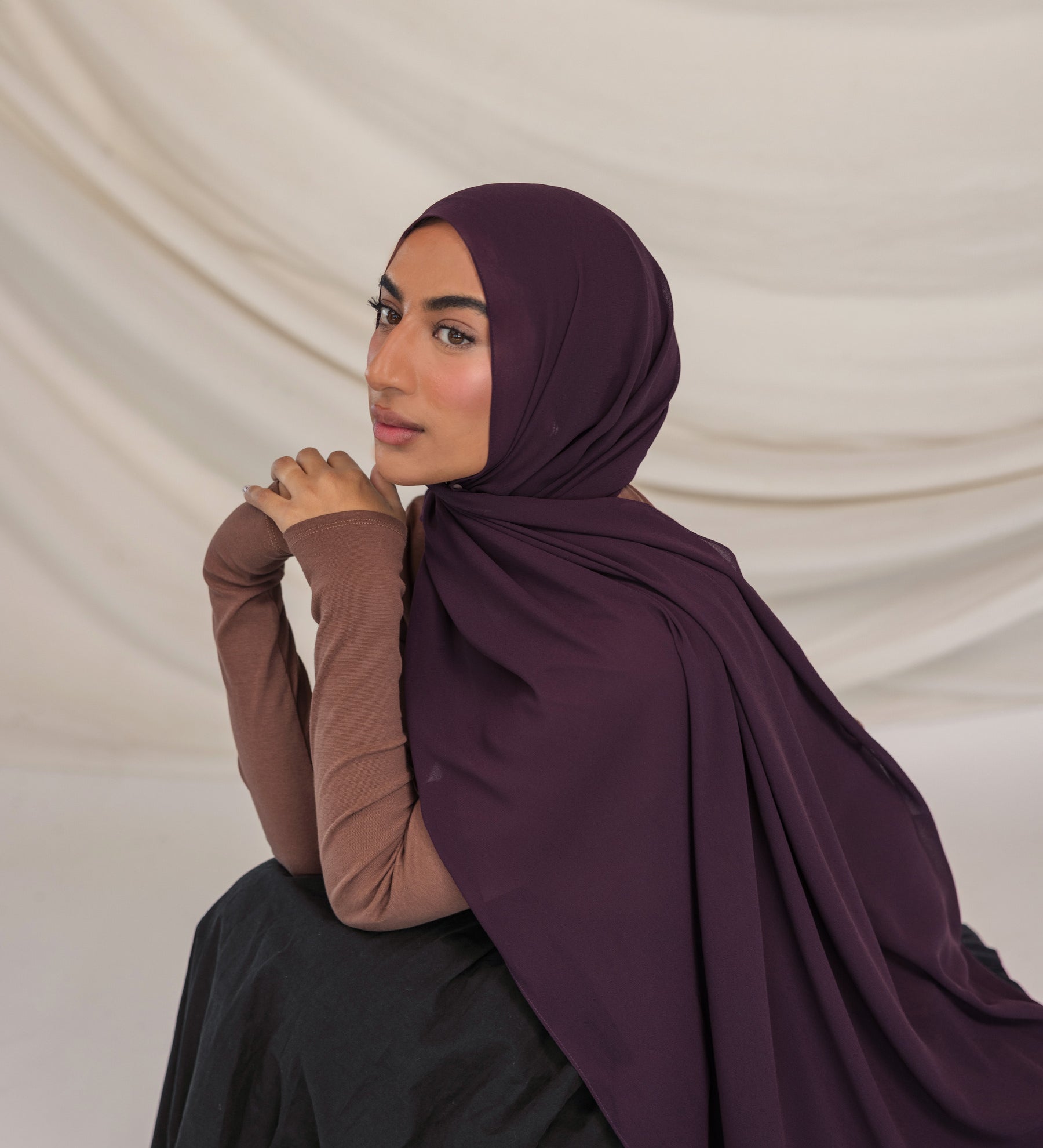 Woman wearing a dark purple hijab and brown long-sleeve top against a beige background