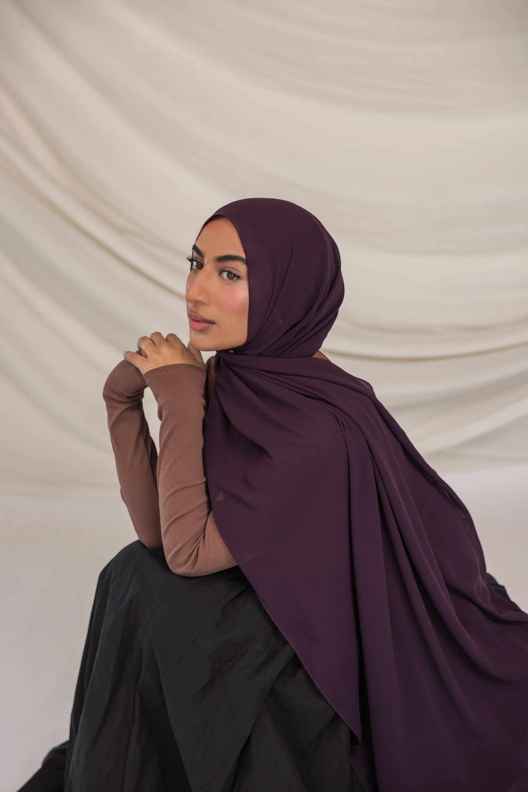 Woman wearing a dark purple hijab and brown long-sleeve top against a beige background
