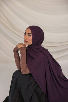 Woman wearing a dark purple hijab and brown long-sleeve top against a beige background
