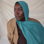 Woman wearing a turquoise hijab and beige coat against a plain background