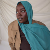 Woman wearing a turquoise hijab and beige coat against a plain background