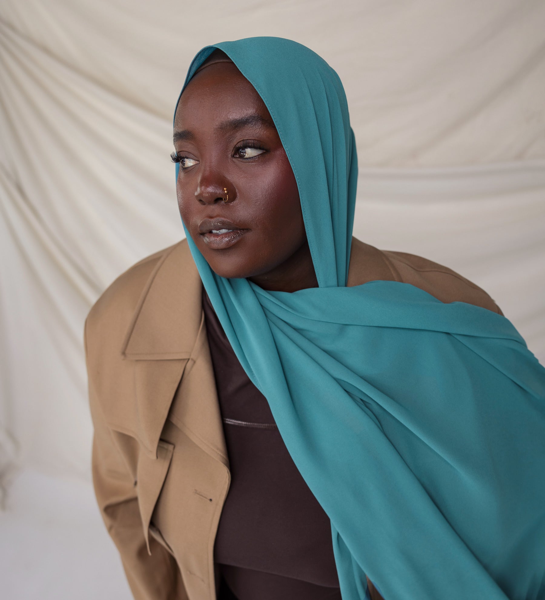 Woman wearing a turquoise hijab and beige coat against a plain background