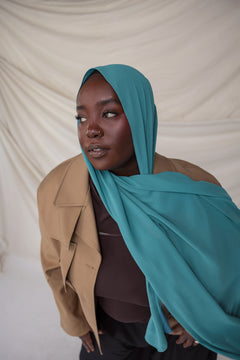 Woman wearing a turquoise hijab and beige coat against a plain background