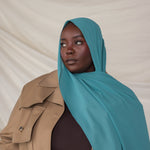 Person wearing a teal hijab and beige trench coat against a plain background