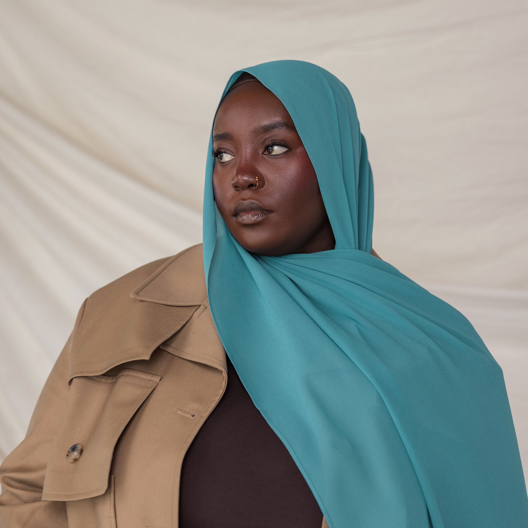 Person wearing a teal hijab and beige trench coat against a plain background