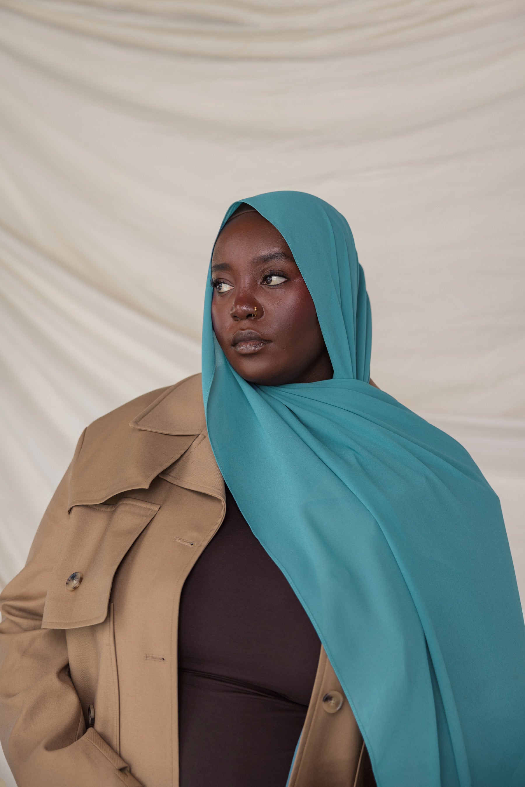 Person wearing a teal hijab and beige trench coat against a plain background