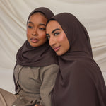 Two women wearing brown hijabs sitting close together against a beige background