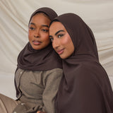 Two women wearing brown hijabs sitting close together against a beige background