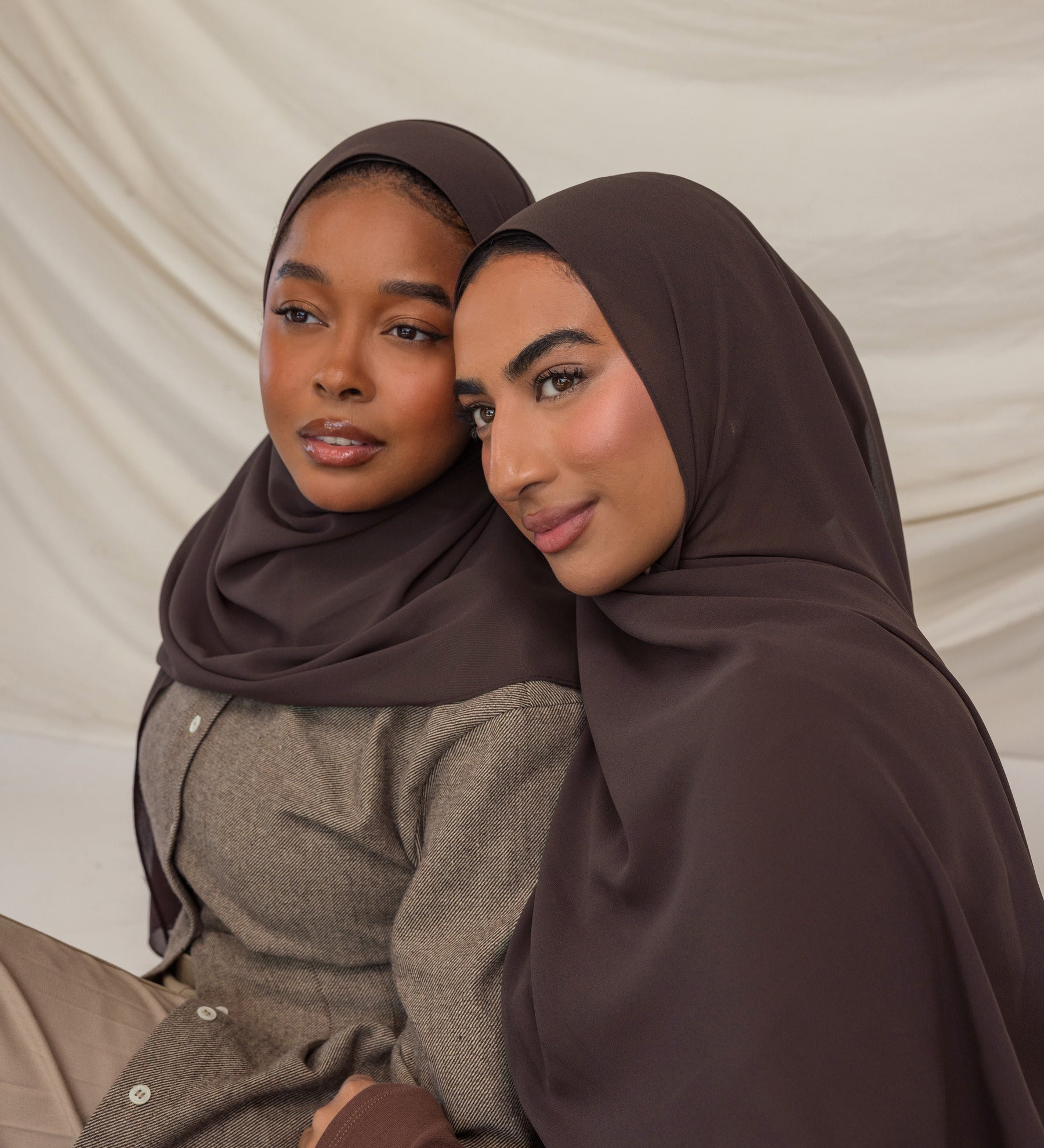 Two women wearing brown hijabs sitting close together against a beige background