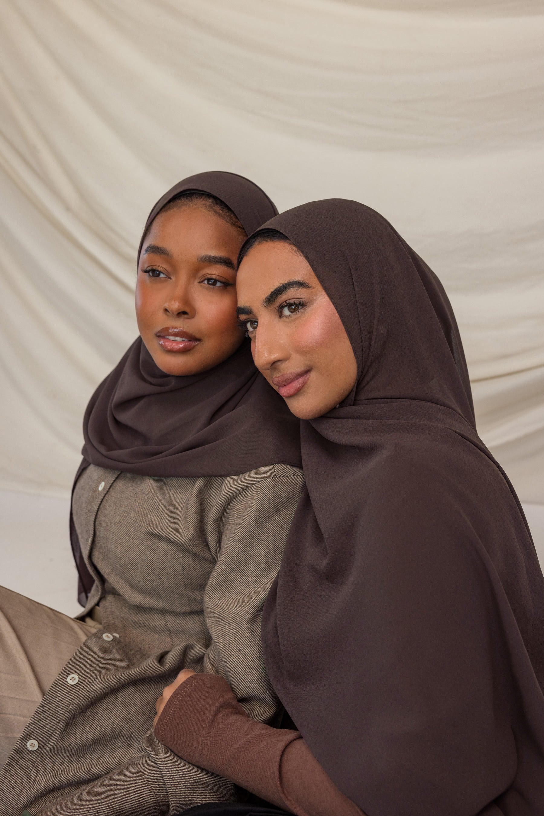 Two women wearing brown hijabs sitting close together against a beige background