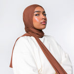 A woman wearing a draped brown hijab and a loose white top, photographed against a clean light gray studio background.