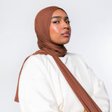 A woman wearing a draped brown hijab and a loose white top, photographed against a clean light gray studio background.