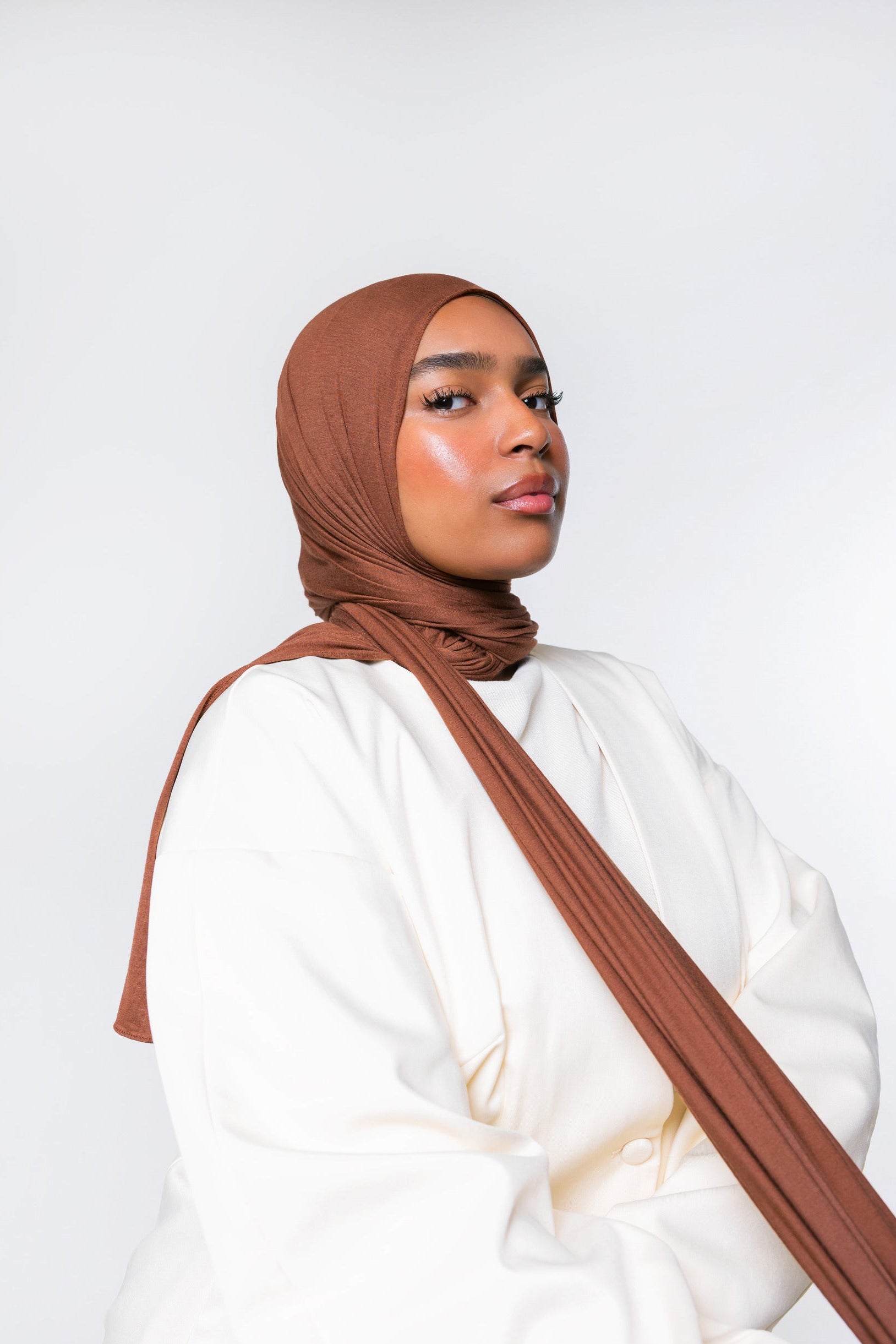 A woman wearing a draped brown hijab and a loose white top, photographed against a clean light gray studio background.