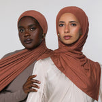 Two women modeling draped terracotta-colored hijabs against a plain light gray studio background. The woman in front wears a cream top, and the woman behind wears a taupe top.