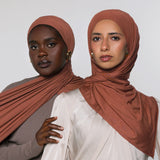 Two women modeling draped terracotta-colored hijabs against a plain light gray studio background. The woman in front wears a cream top, and the woman behind wears a taupe top.