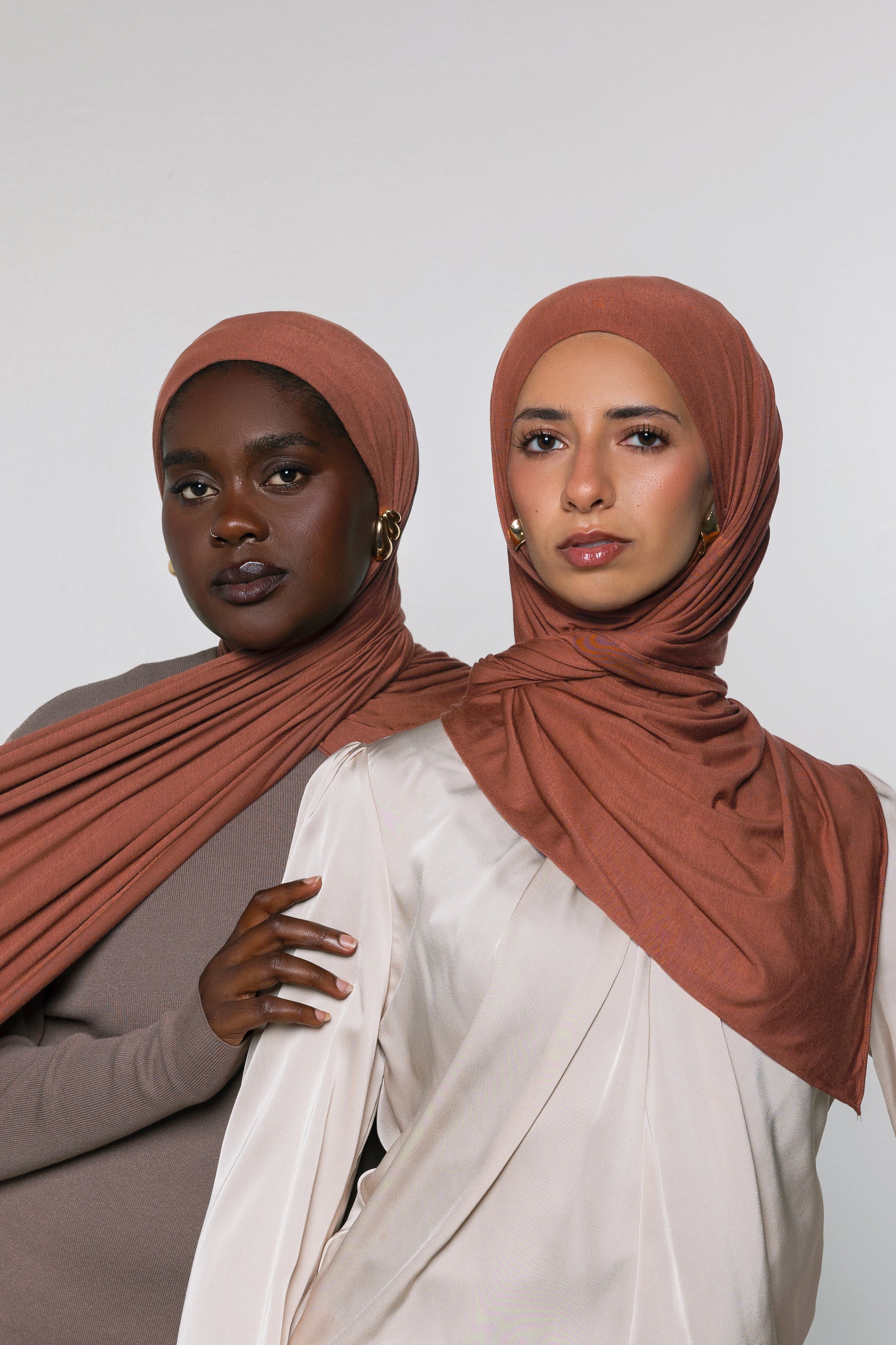 Two women modeling draped terracotta-colored hijabs against a plain light gray studio background. The woman in front wears a cream top, and the woman behind wears a taupe top.