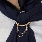 A close-up of a navy blue scarf tied around the neck of a person, secured with a gold-toned ring and layered chain accessory.