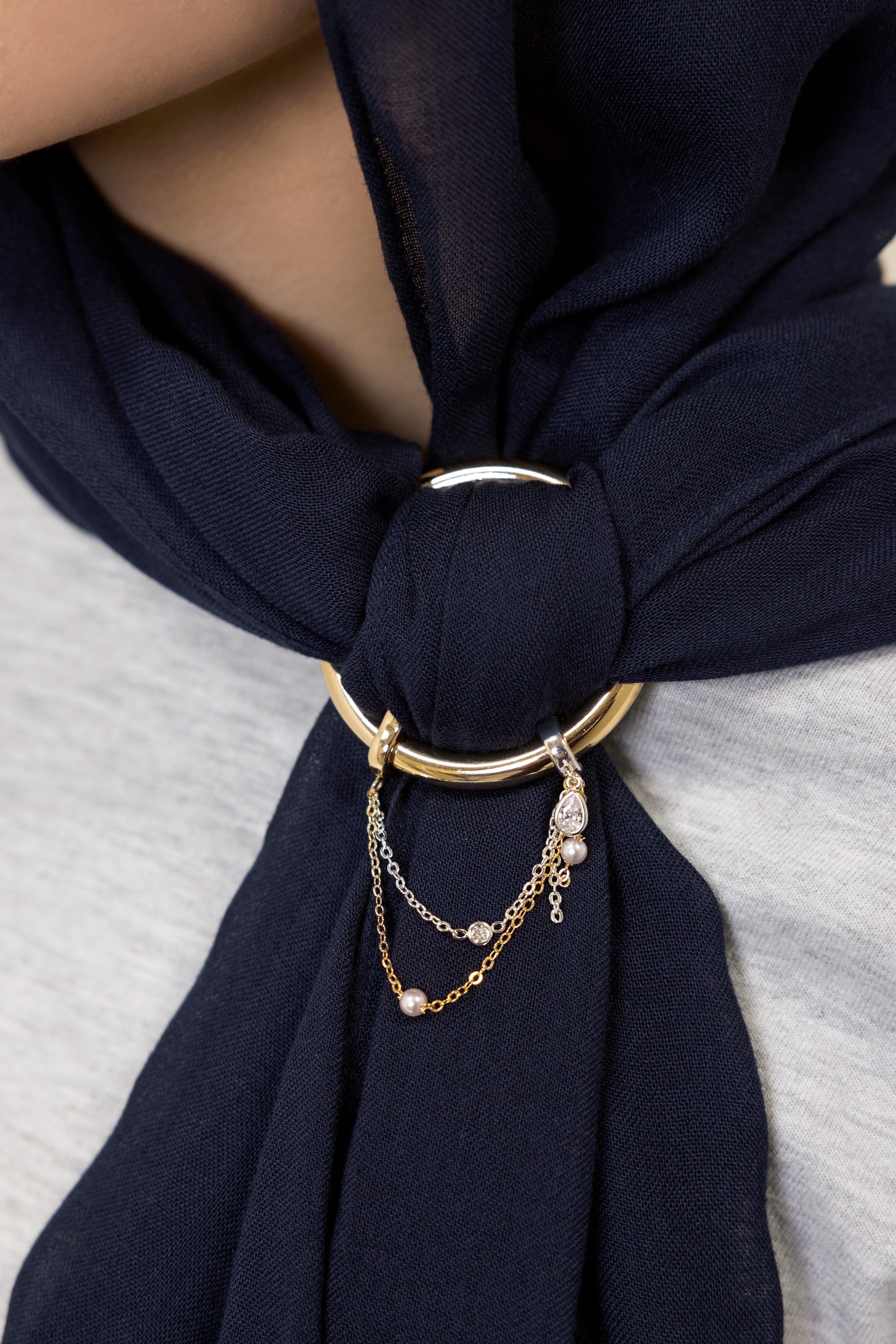 A close-up of a navy blue scarf tied around the neck of a person, secured with a gold-toned ring and layered chain accessory.