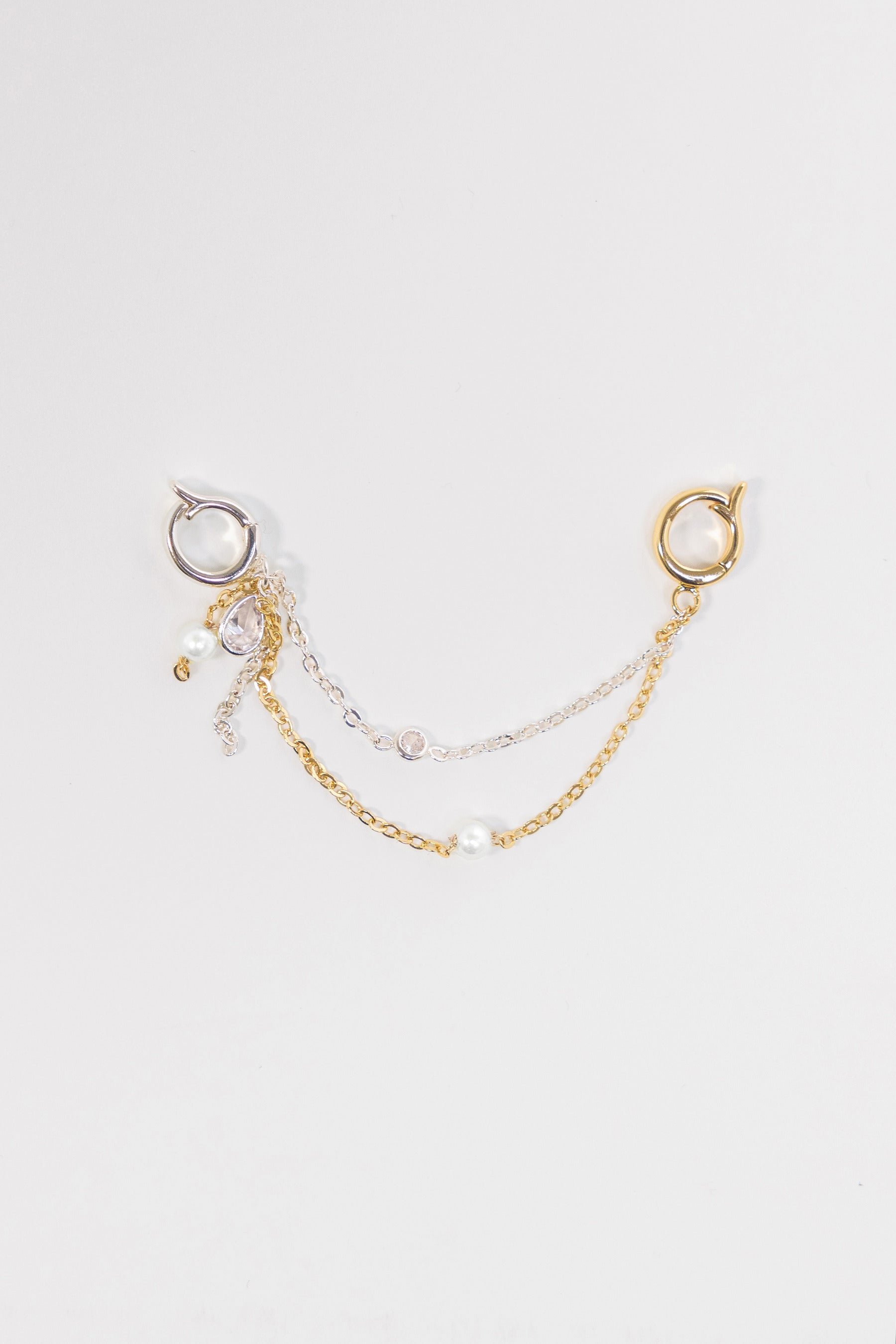 A delicate double-layered chain earring accessory featuring a gold chain with a small pearl bead and a silver chain with a clear rhinestone, attached to a gold and silver hoop earring on a light gray background.