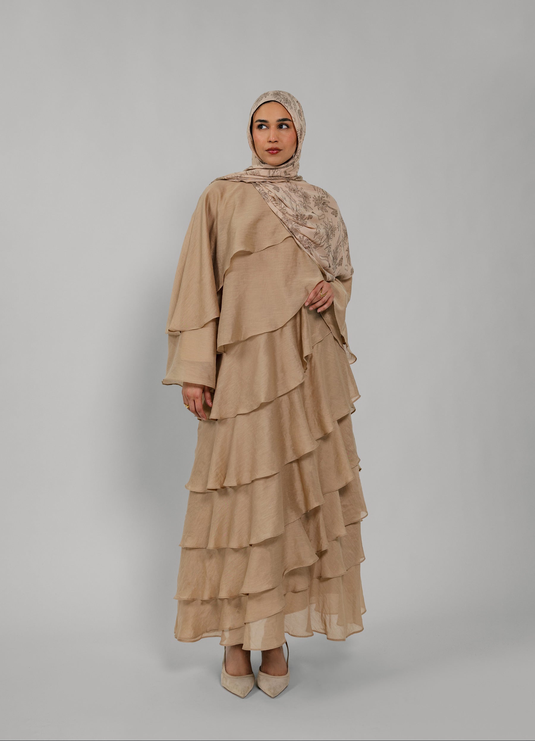 Woman wearing a beige layered dress and hijab on a gray background