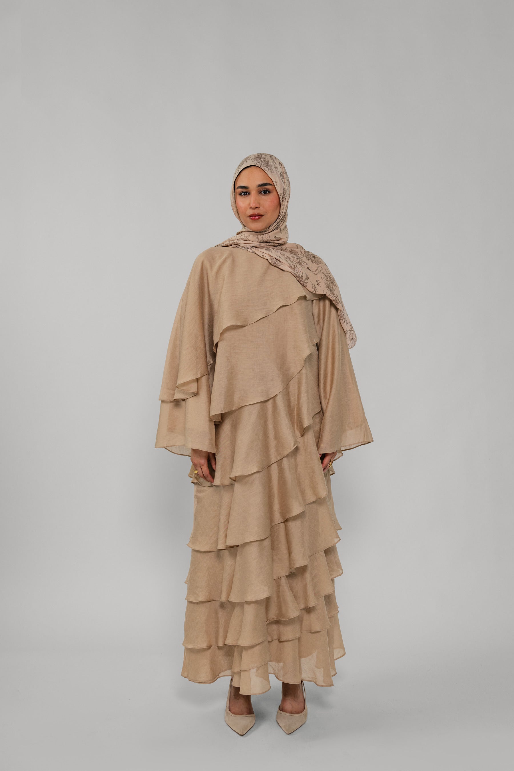 Woman wearing a beige layered dress and hijab on a gray background