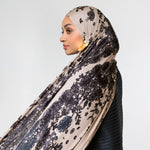 A woman in profile wearing a beige hijab with a black floral lace pattern and a black ribbed top, set against a plain light gray background.