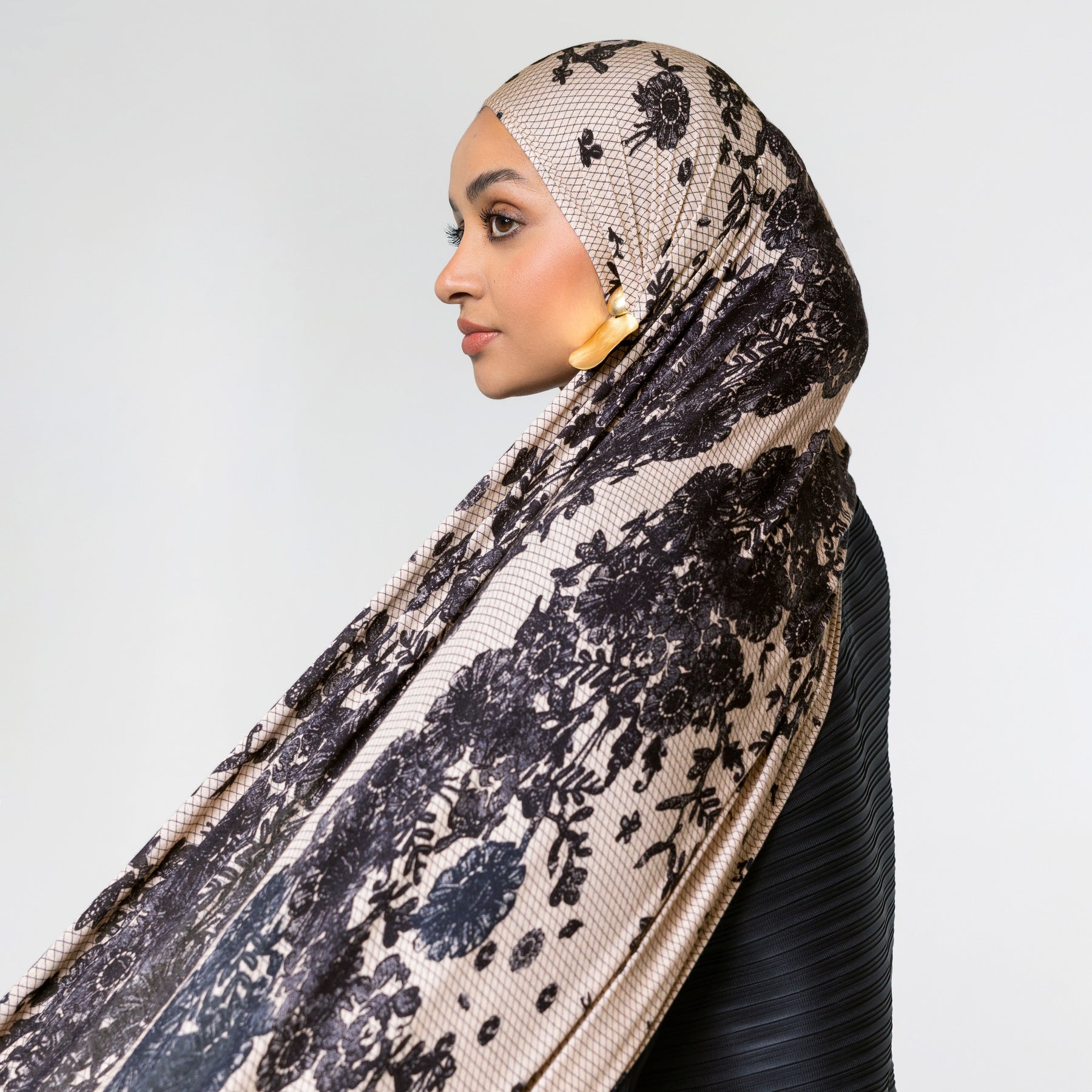 A woman in profile wearing a beige hijab with a black floral lace pattern and a black ribbed top, set against a plain light gray background.