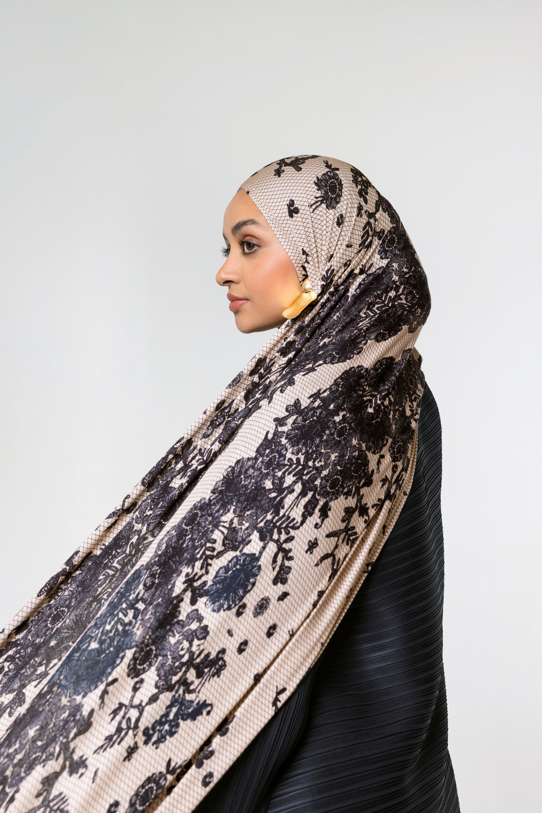 A woman in profile wearing a beige hijab with a black floral lace pattern and a black ribbed top, set against a plain light gray background.