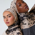 Two women wearing patterned beige and black hijabs, posed closely against a plain light gray studio background.