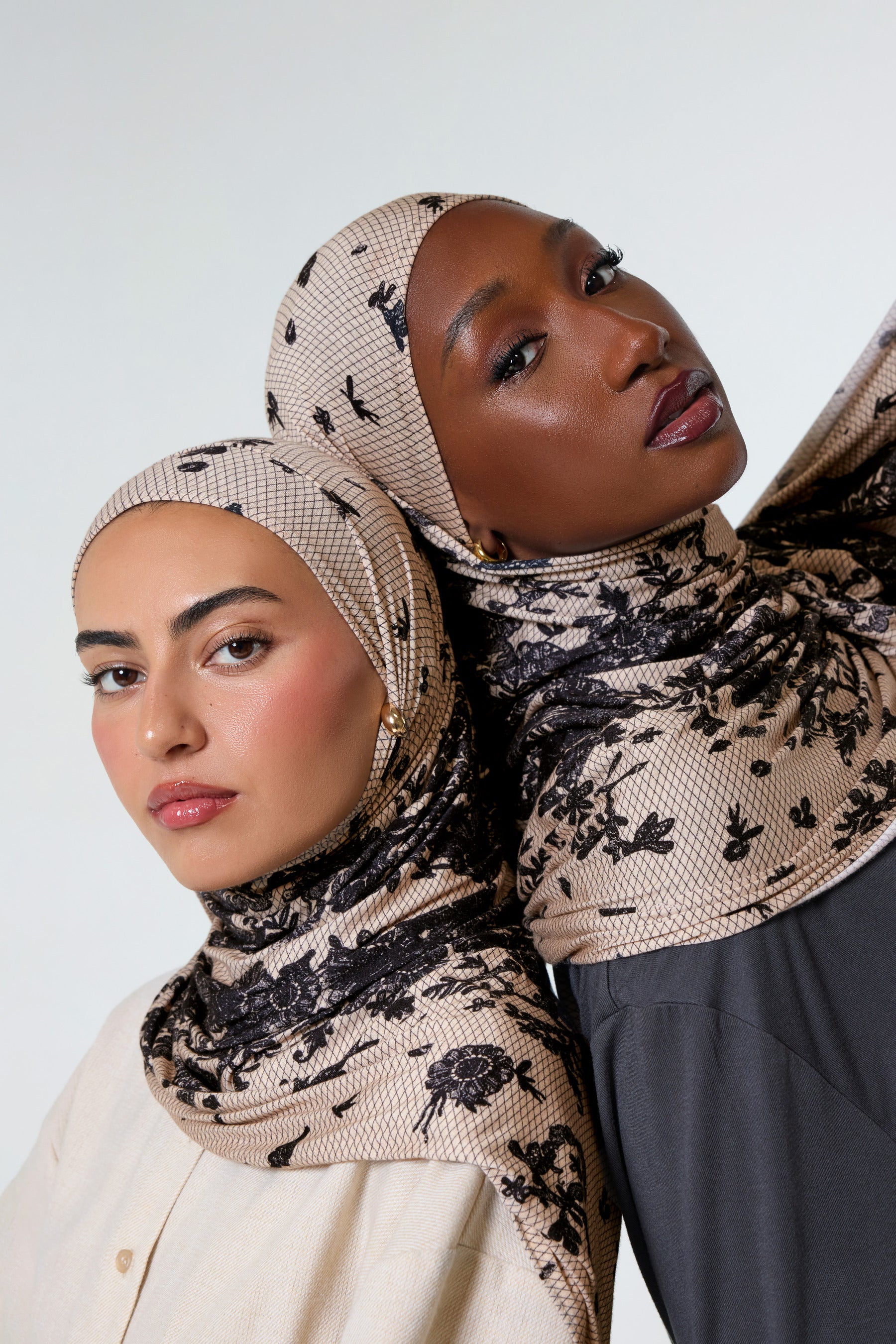 Two women wearing patterned beige and black hijabs, posed closely against a plain light gray studio background.