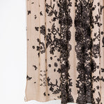 Close-up of a sheer beige mesh fabric with vertical black floral lace panels and scattered black floral motifs, shown against a plain white background.