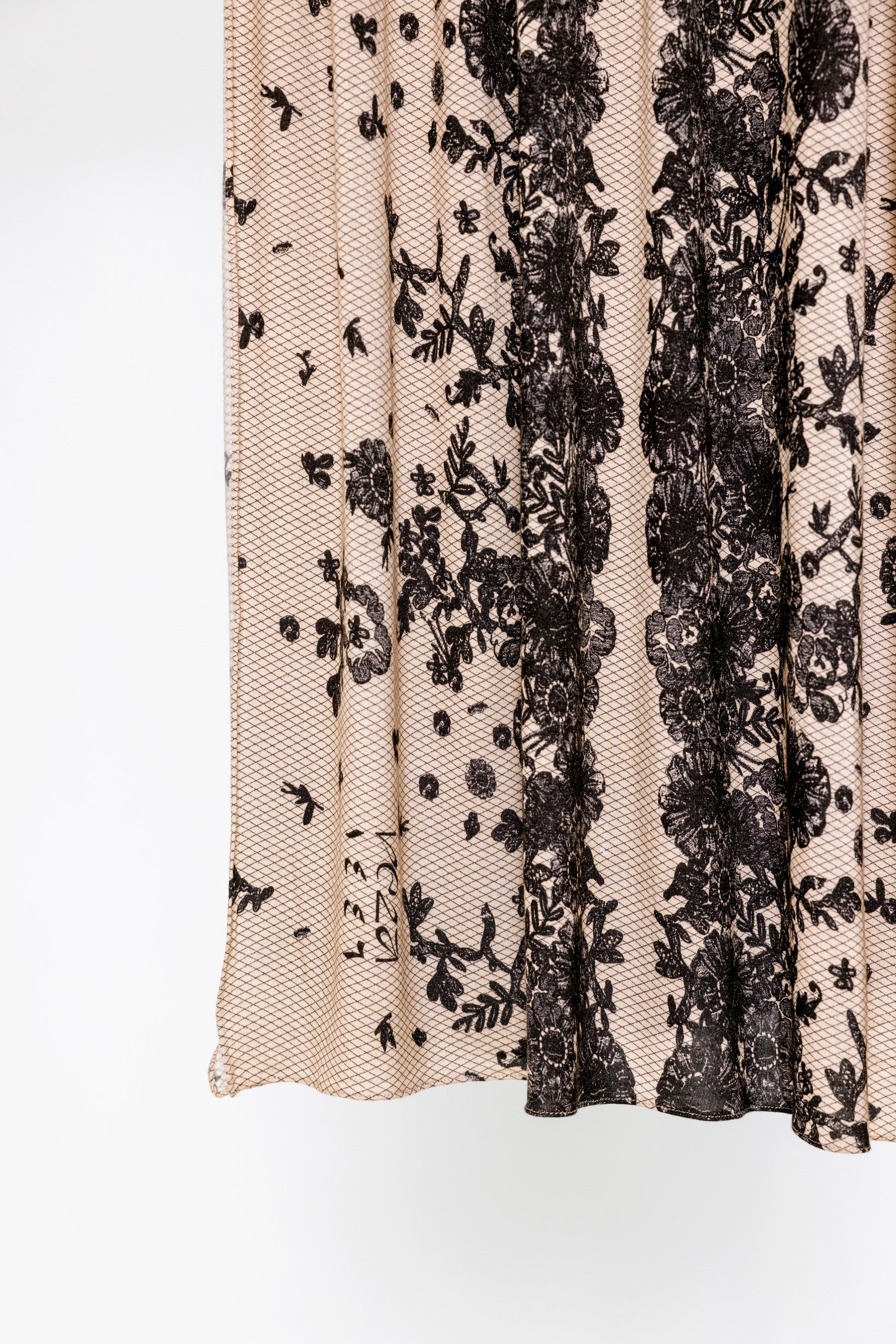 Close-up of a sheer beige mesh fabric with vertical black floral lace panels and scattered black floral motifs, shown against a plain white background.