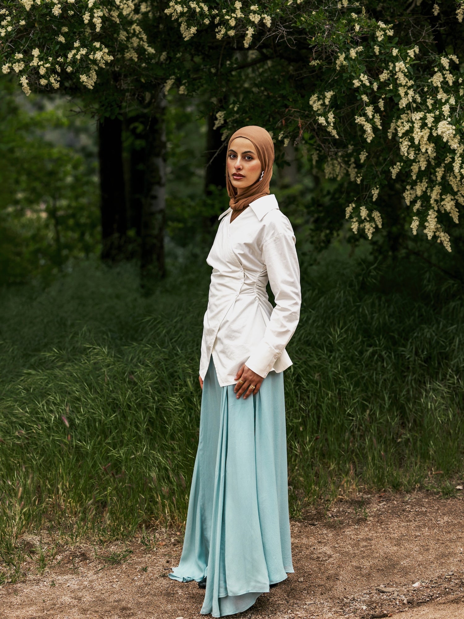 Woman in a white shirt and light blue skirt standing in a forested area with trees and greenery.