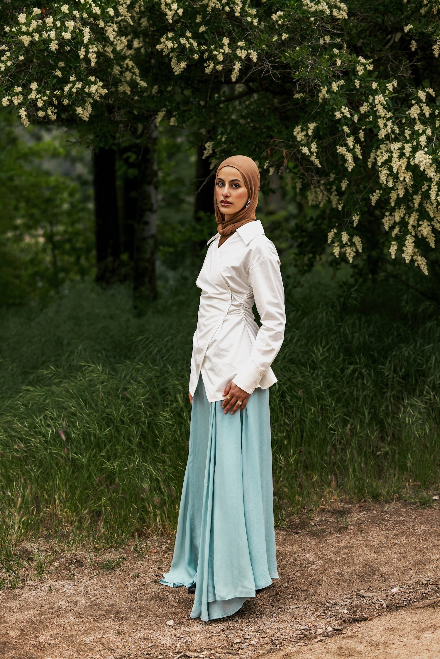 Woman in a white shirt and light blue skirt standing in a forested area with trees and greenery.