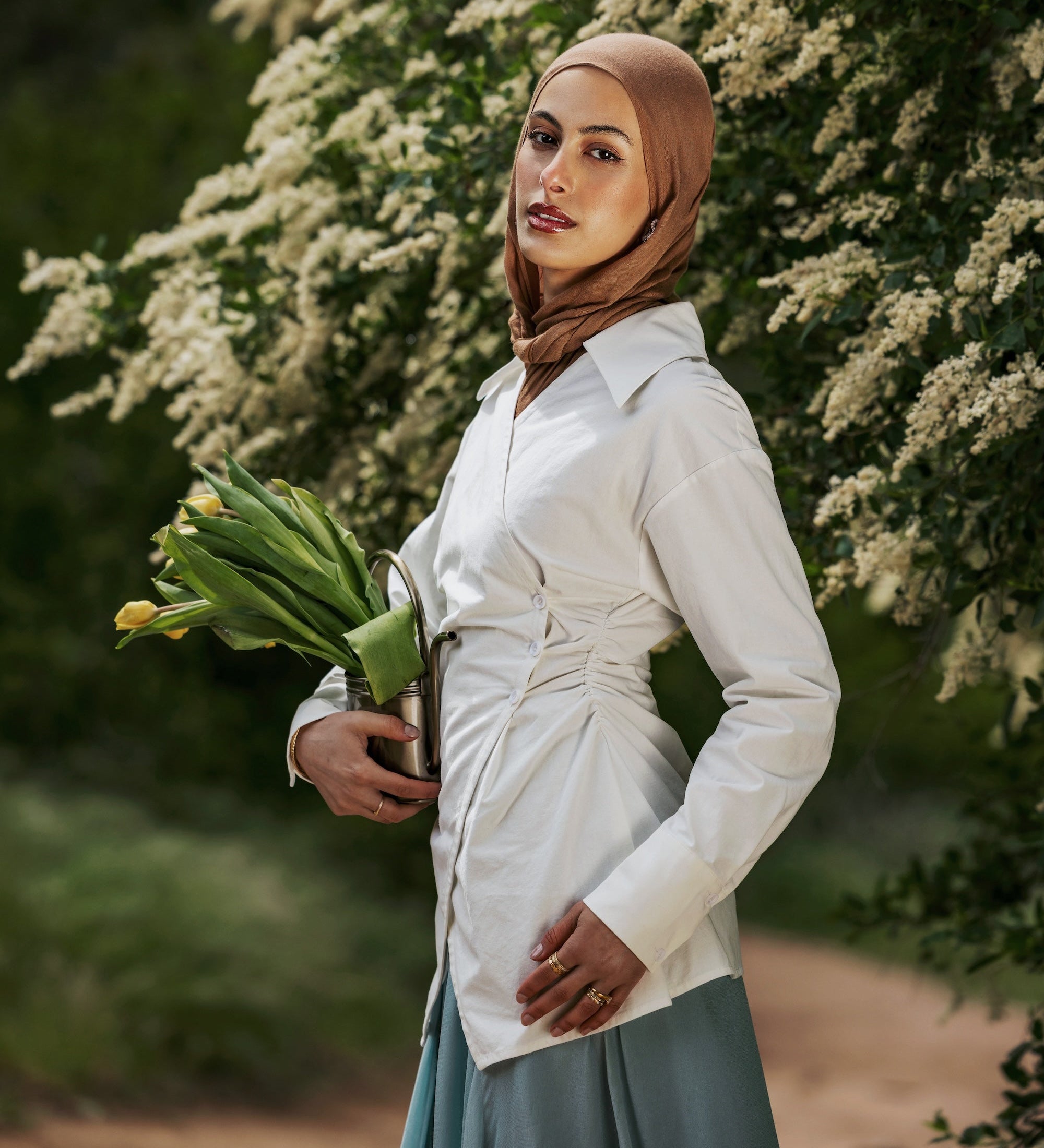 Woman in a hijab holding flowers outdoors with greenery in the background
