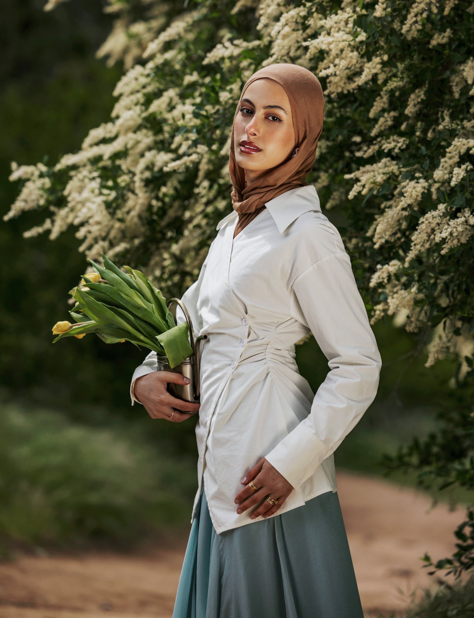 Woman in a hijab holding flowers outdoors with greenery in the background