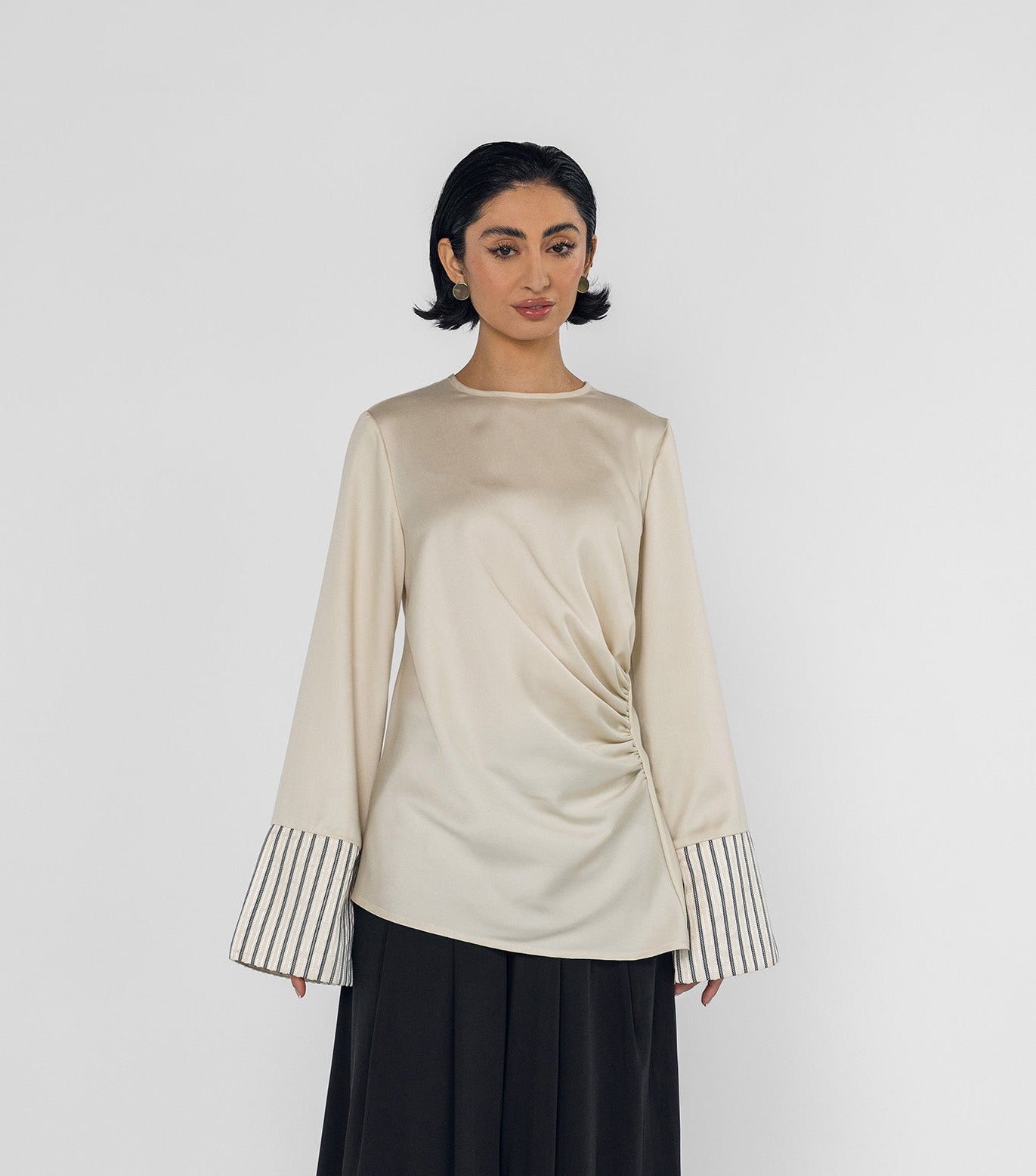 Woman wearing a beige long-sleeve top with striped cuffs and black wide-leg pants on a white background