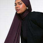 A woman wearing a deep plum-colored hijab and a black blazer, photographed against a clean light gray studio background.