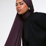A woman wearing a deep plum-colored hijab and a black blazer, photographed against a clean light gray studio background.