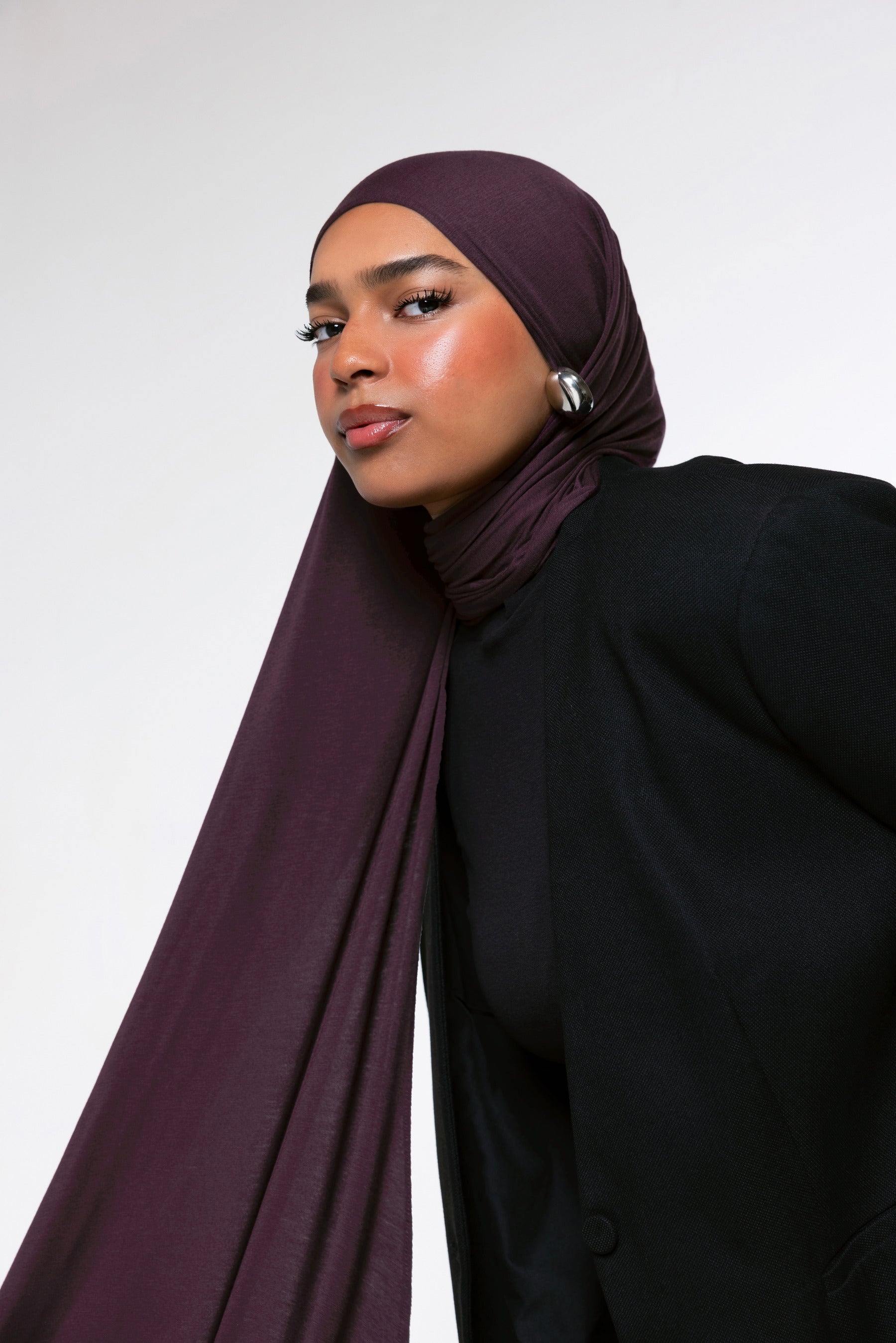A woman wearing a deep plum-colored hijab and a black blazer, photographed against a clean light gray studio background.