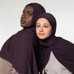 Two women wearing matching deep plum purple hijabs, posed against a plain light gray studio background.