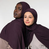 Two women wearing matching deep plum purple hijabs, posed against a plain light gray studio background.