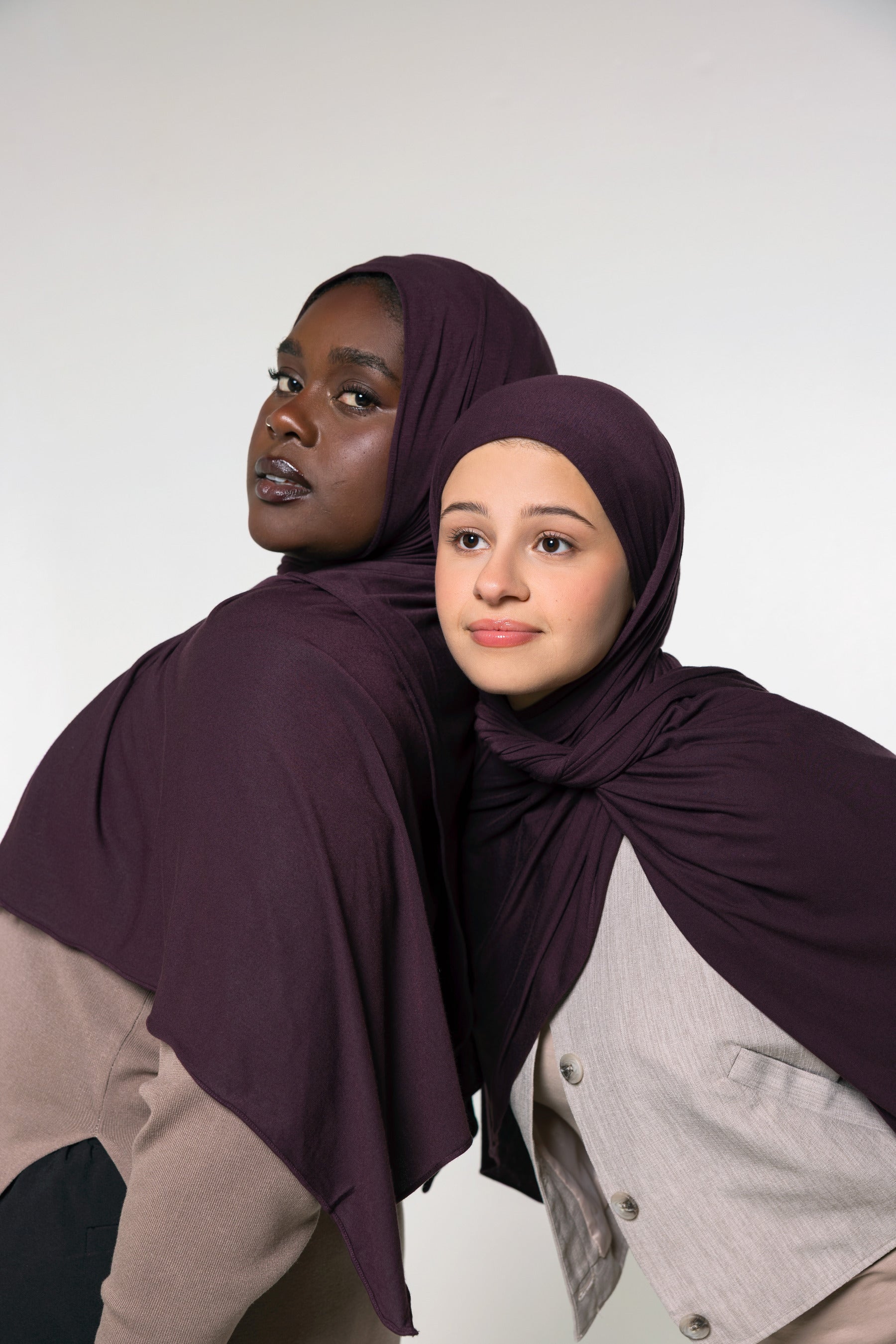 Two women wearing matching deep plum purple hijabs, posed against a plain light gray studio background.