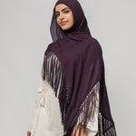 A woman wearing a deep plum purple hijab with long fringe trim draped over her shoulders, styled over a white top and black pants, against a solid light gray studio background.