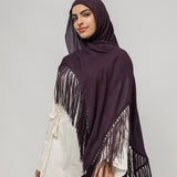 A woman wearing a deep plum purple hijab with long fringe trim draped over her shoulders, styled over a white top and black pants, against a solid light gray studio background.