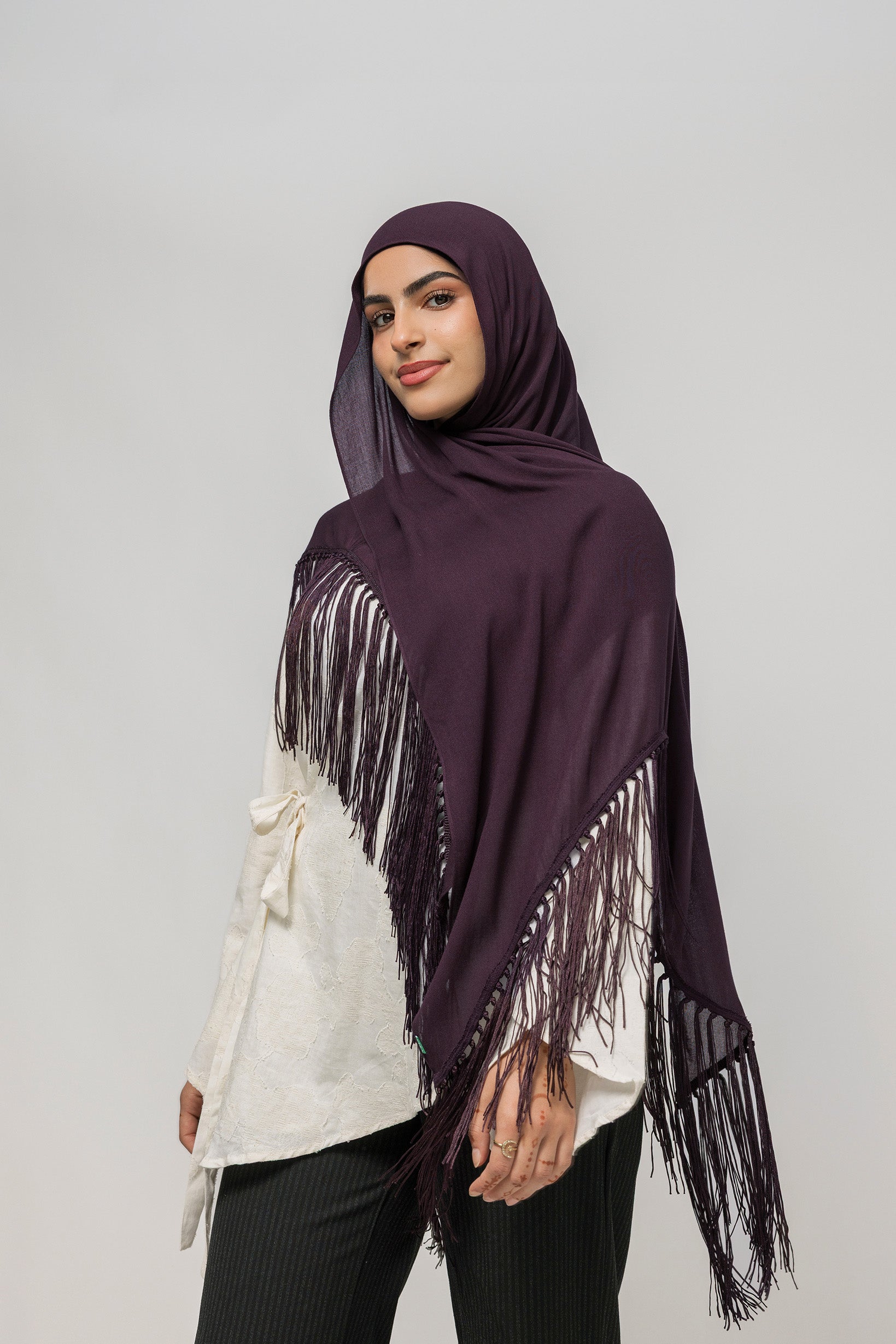 A woman wearing a deep plum purple hijab with long fringe trim draped over her shoulders, styled over a white top and black pants, against a solid light gray studio background.