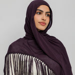 A woman wearing a deep plum purple hijab and a matching fringed shawl over a cream-colored top, photographed against a solid light gray background.