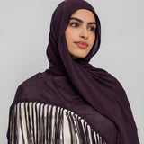 A woman wearing a deep plum purple hijab and a matching fringed shawl over a cream-colored top, photographed against a solid light gray background.