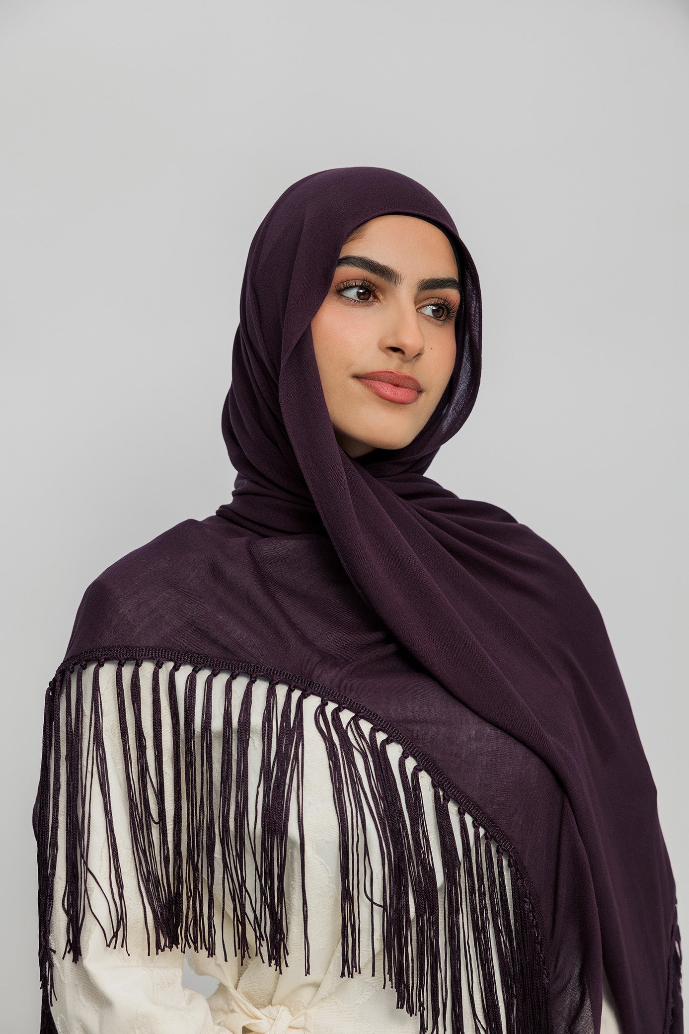 A woman wearing a deep plum purple hijab and a matching fringed shawl over a cream-colored top, photographed against a solid light gray background.